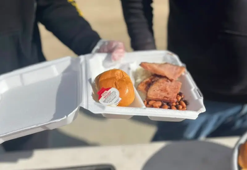 A person holding a takeout container with a sandwich, meat, baked beans, and sauce in a sunny outdoor setting.